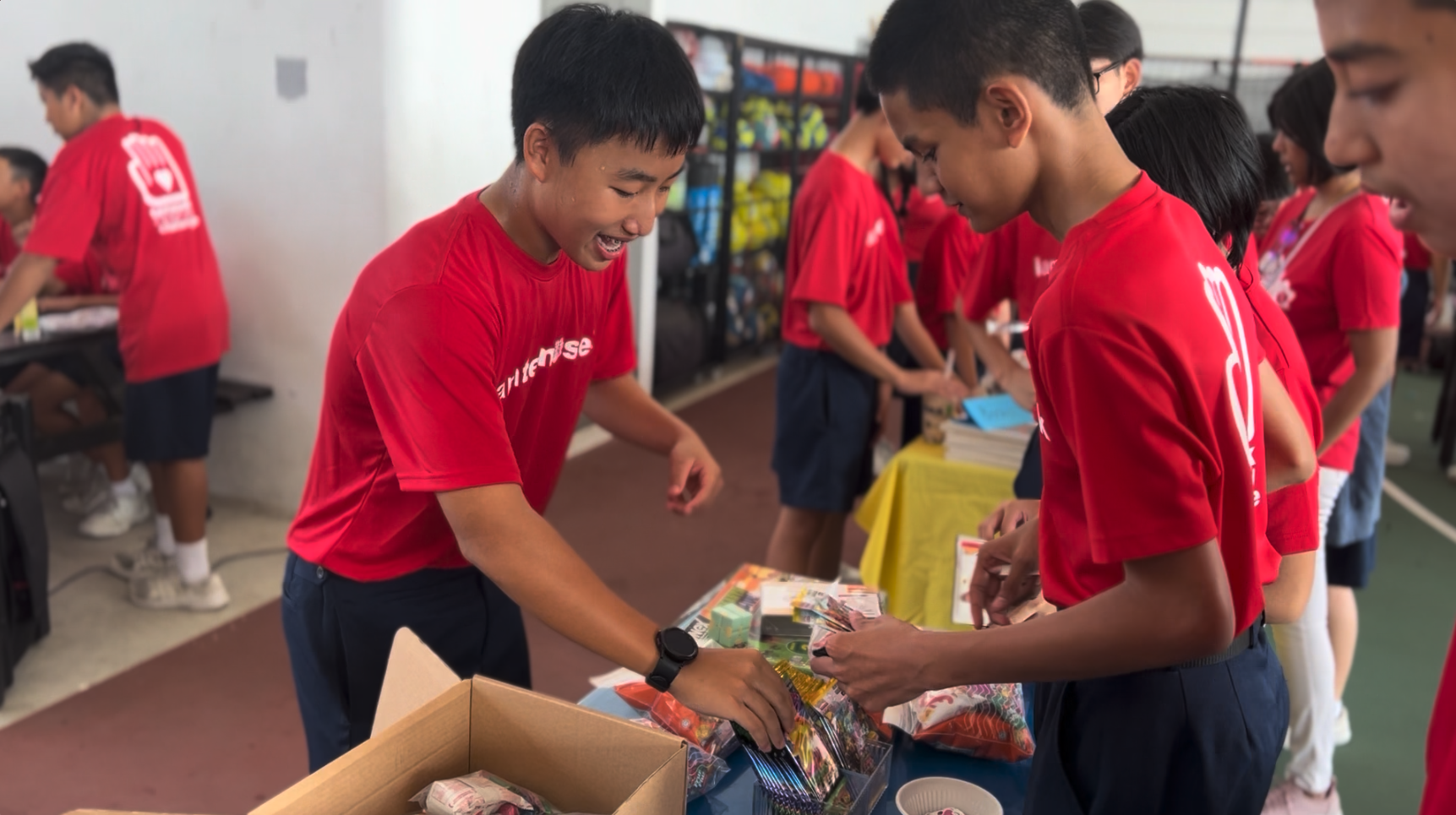 Several youth in matching red shirts sort items from cardboard boxes at tables during a volunteer event.